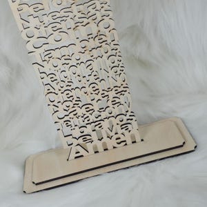 Puede incluir: Un letrero de madera clara con la palabra "Amen" visible. El letrero tiene una base en capas y una secci&oacute;n superior decorativa con letras recortadas. El fondo es una superficie suave, blanca y esponjosa.