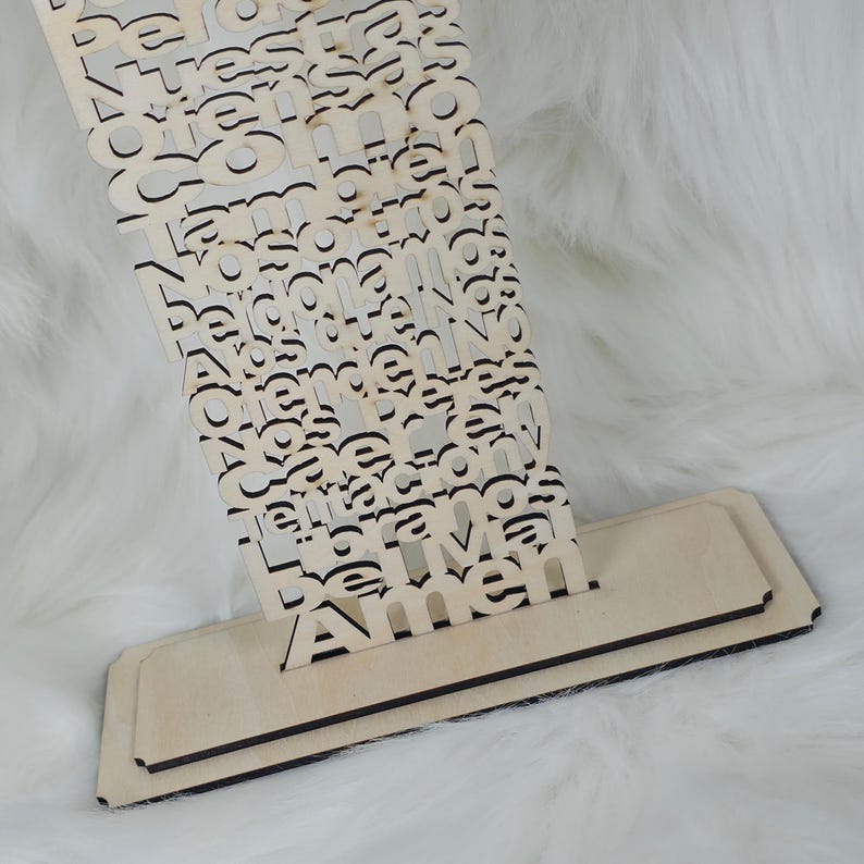 Puede incluir: Un letrero de madera clara con la palabra "Amen" visible. El letrero tiene una base en capas y una secci&oacute;n superior decorativa con letras recortadas. El fondo es una superficie suave, blanca y esponjosa.