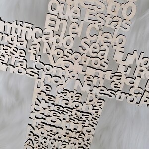 Puede incluir: Una cruz de madera con letras recortadas. La cruz est&aacute; hecha de madera de color claro y presenta texto en una fuente de escritura. El fondo es una superficie suave, blanca y texturizada.