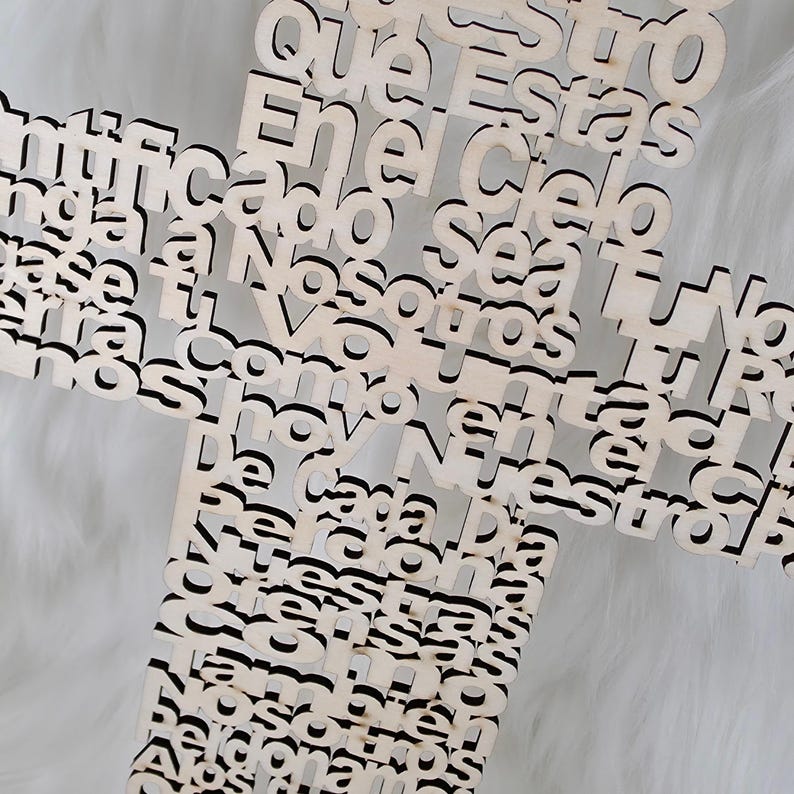 Puede incluir: Una cruz de madera con letras recortadas. La cruz est&aacute; hecha de madera de color claro y presenta texto en una fuente de escritura. El fondo es una superficie suave, blanca y texturizada.