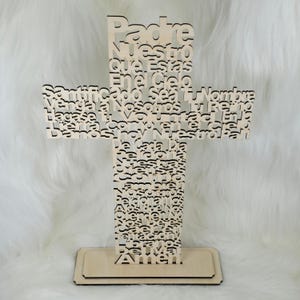 Puede incluir: Una cruz de madera clara con el texto religioso "Padre Nuestro" recortado. La cruz se apoya sobre una base rectangular. El fondo es una superficie blanca y esponjosa.