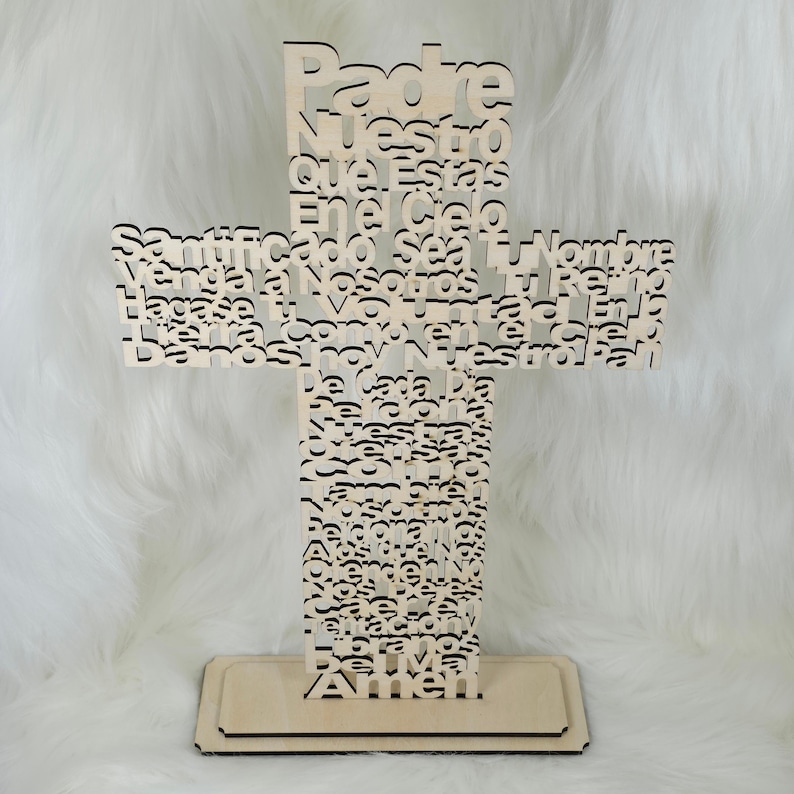 Puede incluir: Una cruz de madera clara con el texto religioso "Padre Nuestro" recortado. La cruz se apoya sobre una base rectangular. El fondo es una superficie blanca y esponjosa.