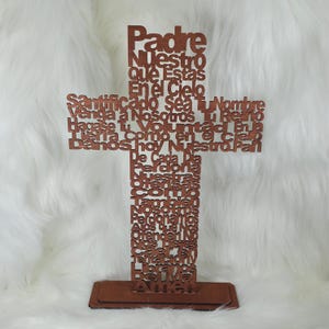 Puede incluir: Una cruz de madera marr&oacute;n con el texto del Padre Nuestro en espa&ntilde;ol. El texto est&aacute; recortado, creando un efecto de capas. La cruz se apoya sobre una base rectangular.