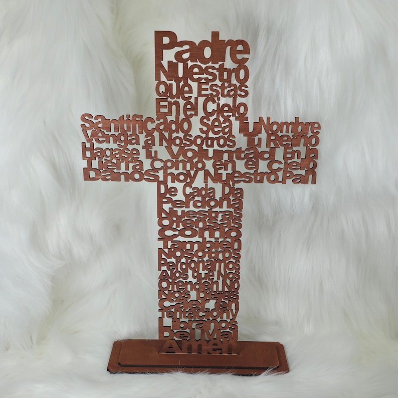Puede incluir: Una cruz de madera marr&oacute;n con el texto del Padre Nuestro en espa&ntilde;ol. El texto est&aacute; recortado, creando un efecto de capas. La cruz se apoya sobre una base rectangular.