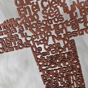 Puede incluir: Una cruz de madera con texto cortado con l&aacute;ser en una fuente de escritura. La cruz es de color marr&oacute;n medio y est&aacute; sobre un fondo blanco texturizado. El texto est&aacute; en espa&ntilde;ol.