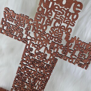 Puede incluir: Una cruz de madera con acabado marr&oacute;n oscuro. La cruz est&aacute; hecha de letras individuales que forman una oraci&oacute;n. La cruz se muestra sobre un fondo blanco y esponjoso.