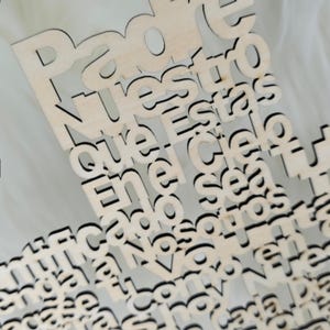 Puede incluir: Primer plano de un recorte de madera con el texto en espa&ntilde;ol "Padre Nuestro que est&aacute;s en el cielo". Las letras est&aacute;n recortadas en madera clara con bordes oscuros, creando un efecto de capas. El fondo es blanco suave y borroso.