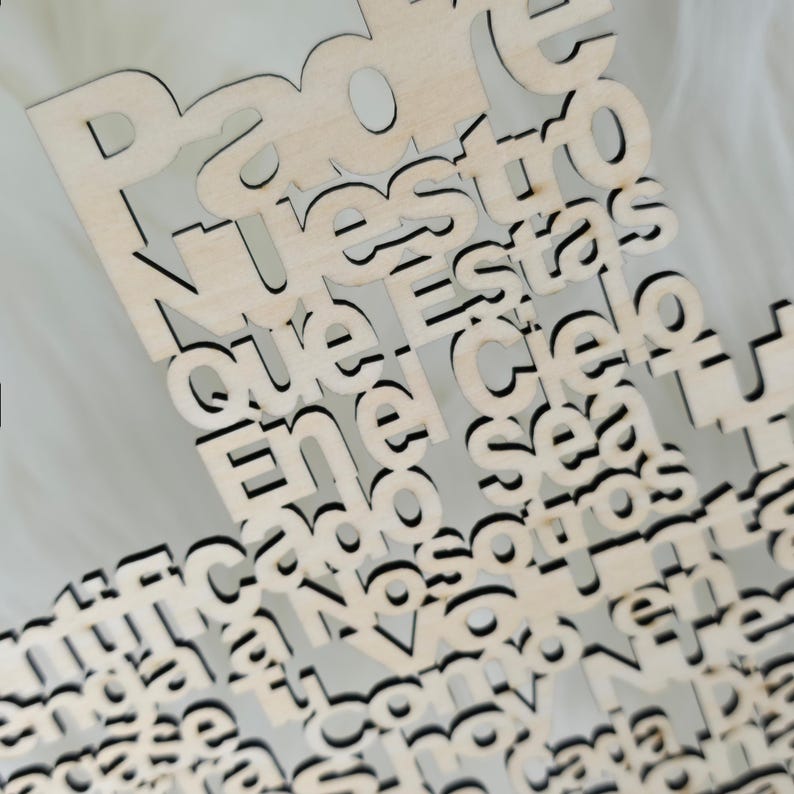 Puede incluir: Primer plano de un recorte de madera con el texto en espa&ntilde;ol "Padre Nuestro que est&aacute;s en el cielo". Las letras est&aacute;n recortadas en madera clara con bordes oscuros, creando un efecto de capas. El fondo es blanco suave y borroso.
