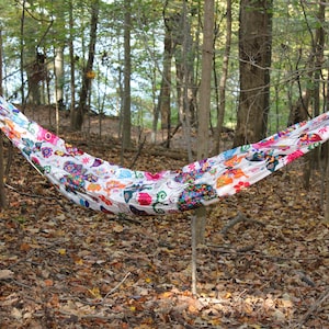 May include: A colorful hammock with a floral pattern hangs between two trees in a wooded area. The hammock is made of a lightweight fabric and is perfect for relaxing outdoors.