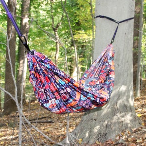 May include: A colorful hammock with a floral pattern hangs between two trees in a forest setting. The hammock is made of a soft, durable fabric and is perfect for relaxing outdoors.