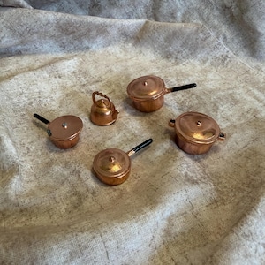 May include: A miniature copper-colored kitchen set consisting of a small kettle, two lidded saucepans with black handles, and a lidded pot with two side handles. Each piece features a metallic finish and is arranged on a textured fabric surface.