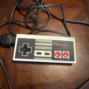 Può includere: Un controller per videogiochi rettangolare grigio e nero con un pad direzionale nero, due pulsanti circolari rossi etichettati A e B e due pulsanti grigi etichettati SELECT e START. Il logo Nintendo è stampato in rosso ed è presente un cavo nero.