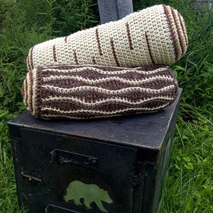 May include: Two crocheted log-shaped pillows, one brown and white, the other white with brown stripes, stacked on top of a black metal box with a bear silhouette painted on it.