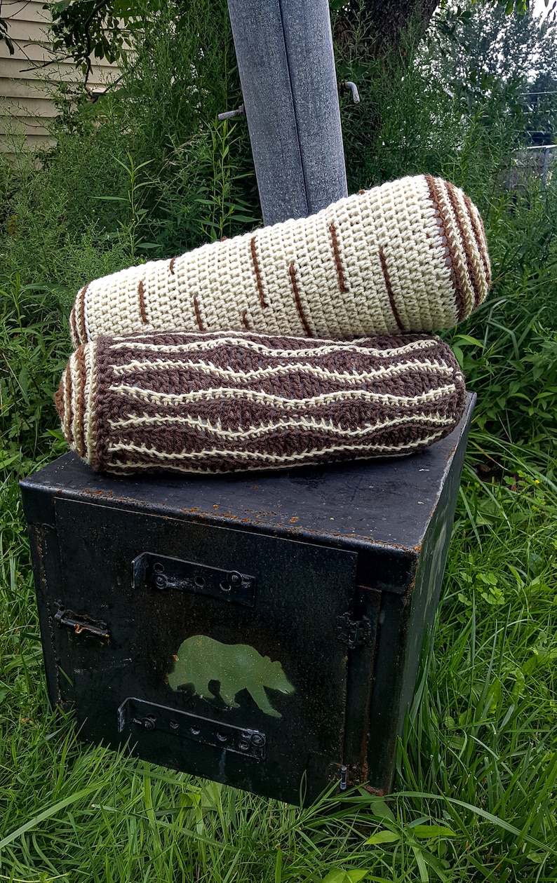 May include: Two crocheted log-shaped pillows, one brown and white, the other white with brown stripes, stacked on top of a black metal box with a bear silhouette painted on it.