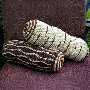 May include: Two crocheted log-shaped pillows, one brown and white striped and the other white with brown stripes, are lying on a purple patterned fabric.