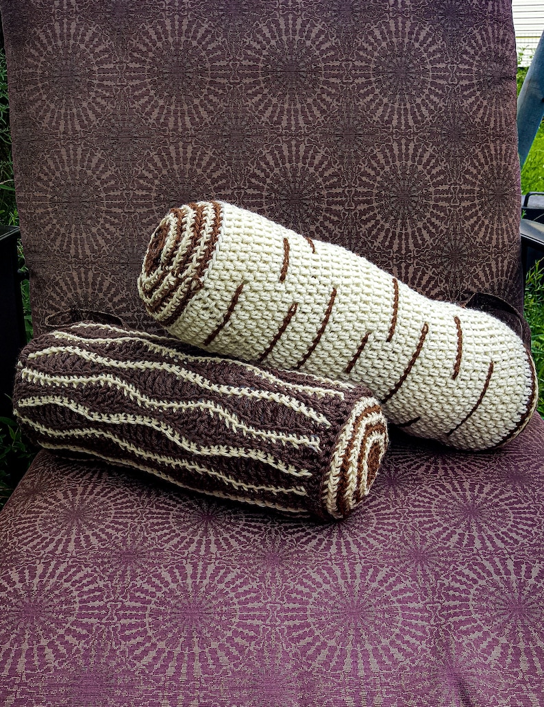 May include: Two crocheted log-shaped pillows, one brown and white striped and the other white with brown stripes, are lying on a purple patterned fabric.