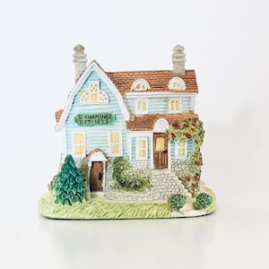May include: A miniature house figurine with a light blue exterior, brown roof, and a green sign that reads "C. Cummings EST 1873." The house has a stone facade, a small tree, and climbing vines. The figurine is on a green base.