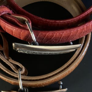 May include: Close-up of three leather belts in shades of red and brown. The belts have silver-coloured rectangular buckles. The leather has a textured, embossed pattern. The belts are arranged in a circular fashion against a dark background.