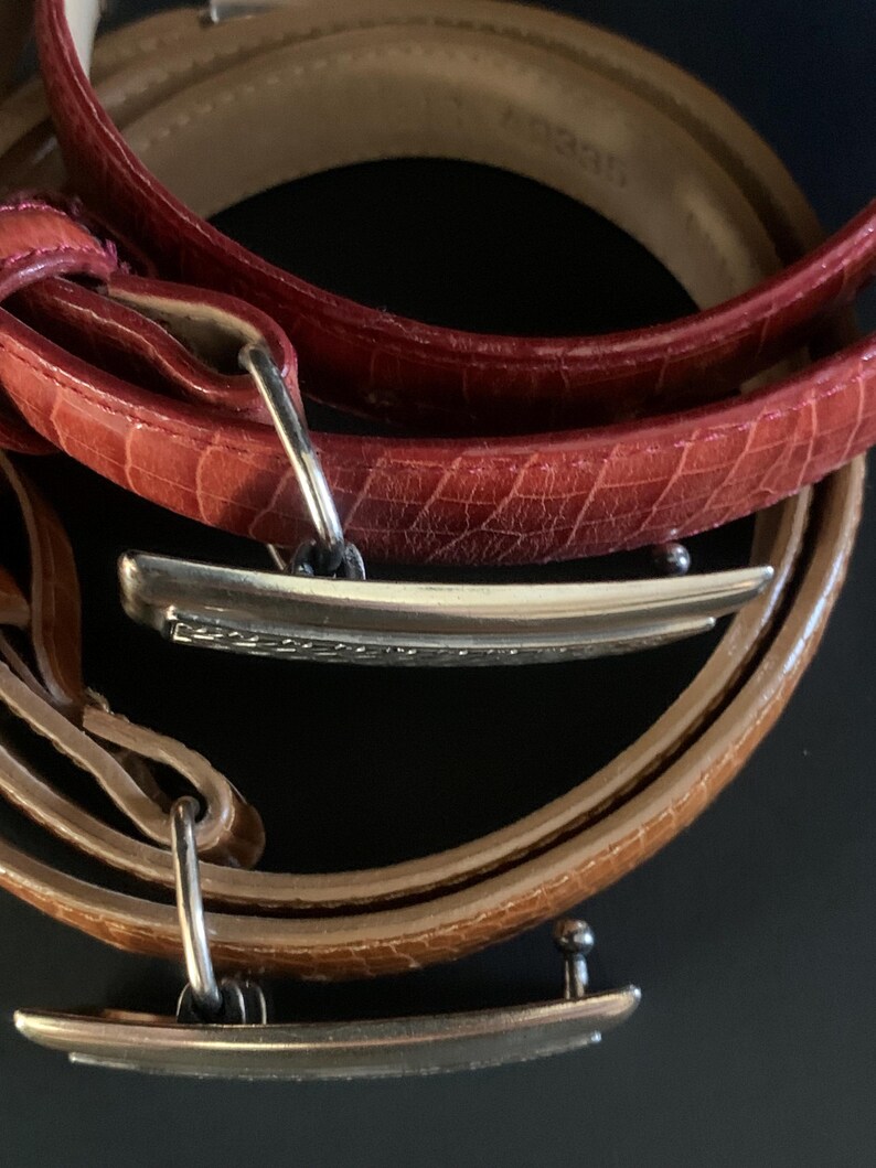May include: Close-up of three leather belts in shades of red and brown. The belts have silver-coloured rectangular buckles. The leather has a textured, embossed pattern. The belts are arranged in a circular fashion against a dark background.