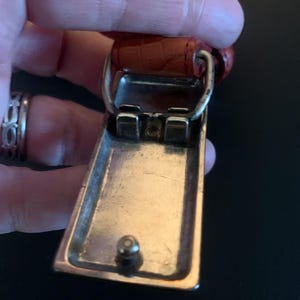 May include: Close-up of a belt buckle with a rectangular, silver-coloured frame and a small, round stud. The buckle is attached to a reddish-brown leather belt with a textured surface. The buckle is held by a hand.