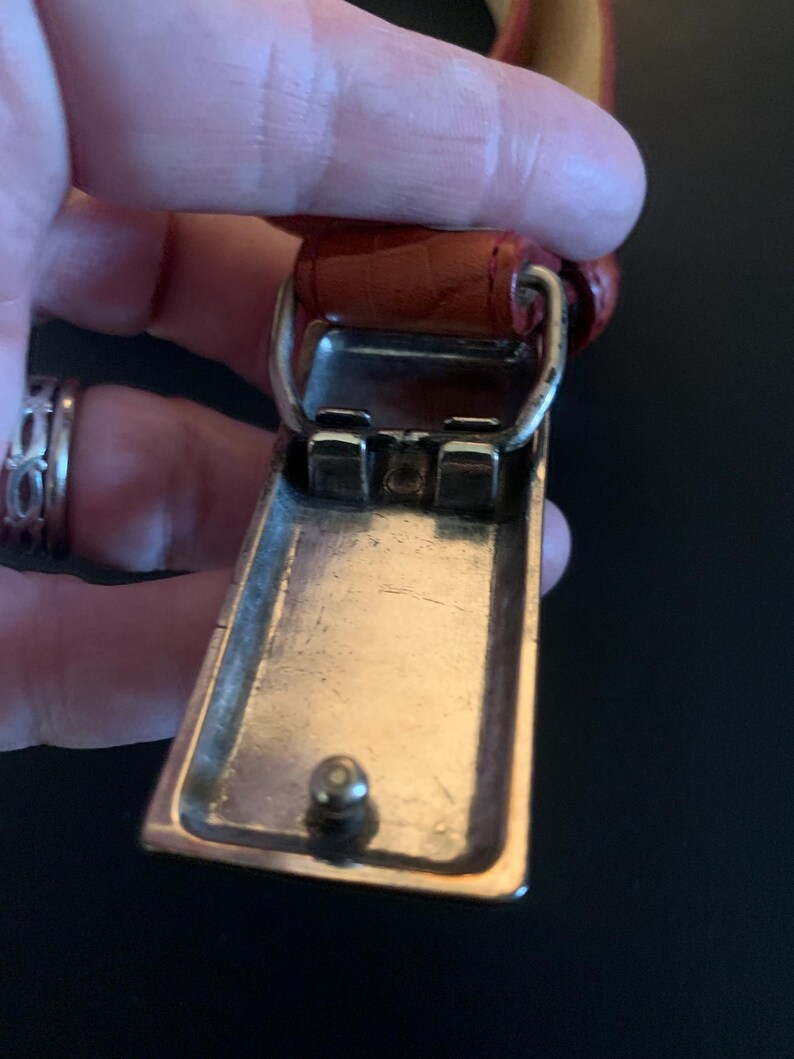 May include: Close-up of a belt buckle with a rectangular, silver-coloured frame and a small, round stud. The buckle is attached to a reddish-brown leather belt with a textured surface. The buckle is held by a hand.