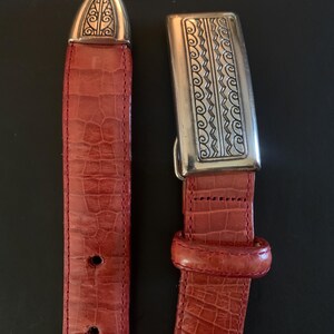 May include: A red leather belt with a silver buckle and tip. The belt has a textured, crocodile-like pattern. The buckle and tip feature ornate silver designs. The belt is shown against a black background.