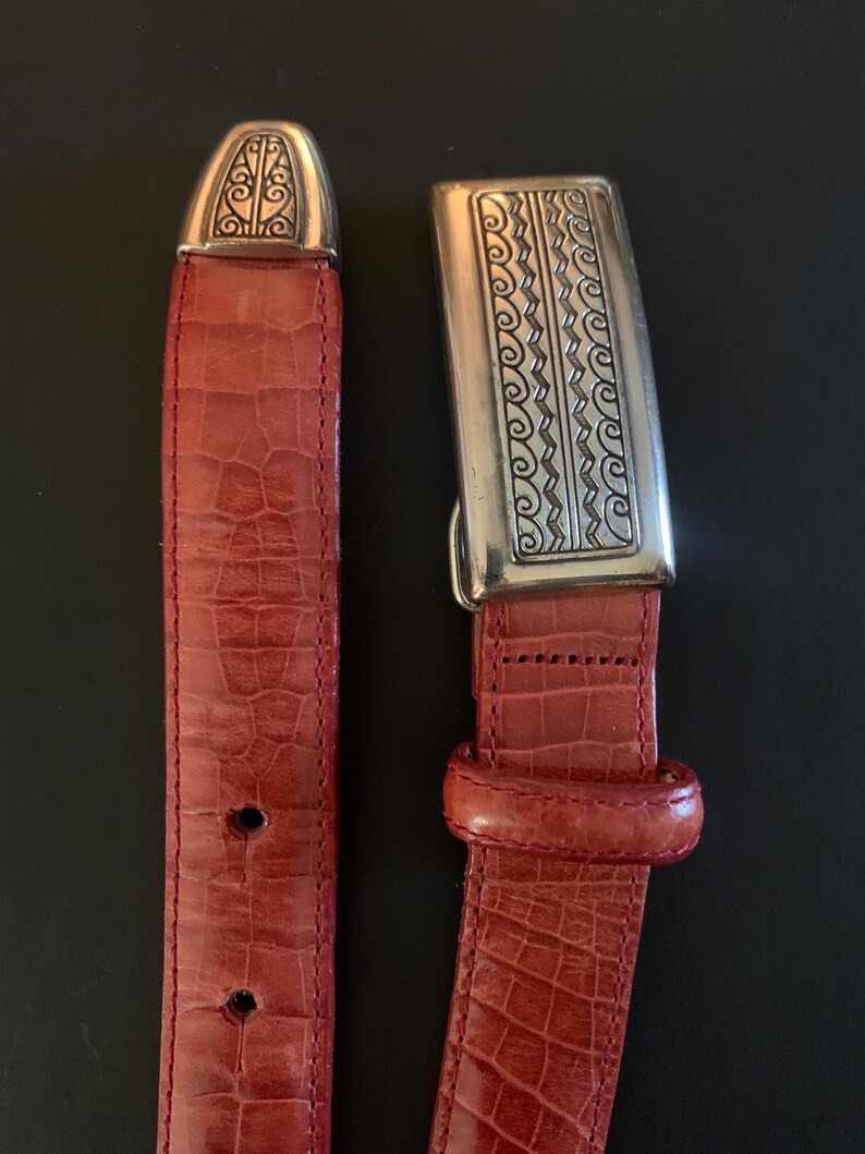 May include: A red leather belt with a silver buckle and tip. The belt has a textured, crocodile-like pattern. The buckle and tip feature ornate silver designs. The belt is shown against a black background.