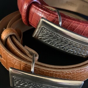 May include: Two leather belts with silver buckles. One is a reddish-orange colour, the other a light brown. The rectangular buckles have an engraved design. The belts have a textured, crocodile-like pattern.
