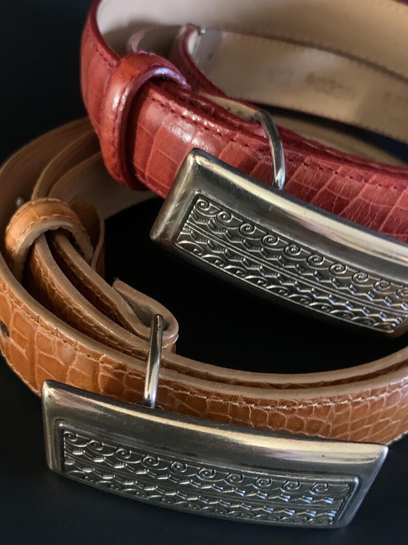 May include: Two leather belts with silver buckles. One is a reddish-orange colour, the other a light brown. The rectangular buckles have an engraved design. The belts have a textured, crocodile-like pattern.
