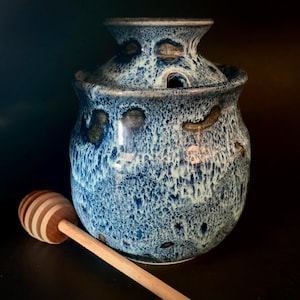 May include: A blue ceramic honey pot with a lid, featuring a speckled glaze and a wooden honey dipper. The pot has a rounded shape with a small opening in the lid. The dipper is light brown with alternating light and dark wood stripes.