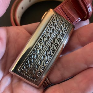 May include: A red leather belt with a rectangular silver buckle. The buckle has an ornate, engraved design with swirling and geometric patterns. The belt is reddish-brown and appears to be made of textured leather.