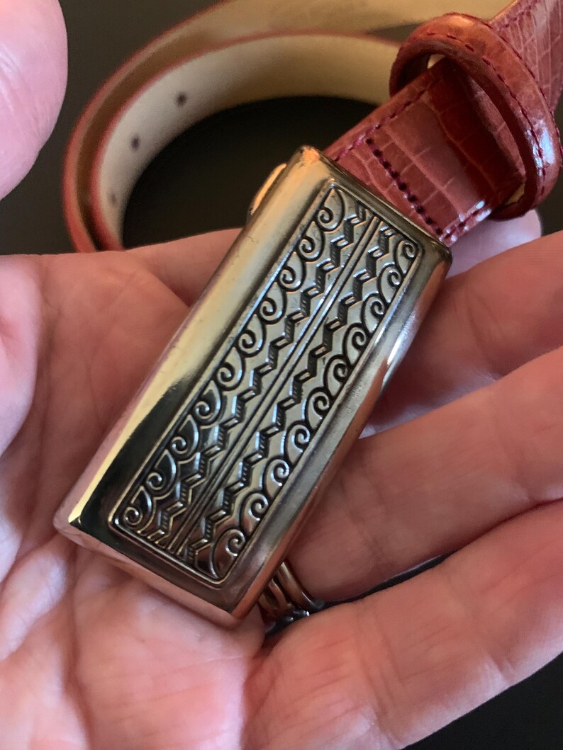 May include: A red leather belt with a rectangular silver buckle. The buckle has an ornate, engraved design with swirling and geometric patterns. The belt is reddish-brown and appears to be made of textured leather.