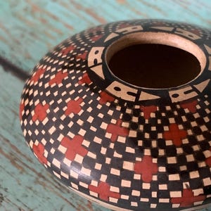 May include: A handcrafted, round pottery vessel with a wide opening. The exterior features a repeating pattern of red crosses, black squares, and cream-colored squares. The rim is black with white geometric shapes.