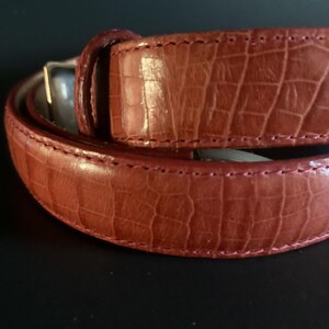 May include: A close-up shot of a brown leather belt with a crocodile skin effect. The belt has a silver buckle and is stitched with a contrasting thread. The leather is a reddish-brown colour.