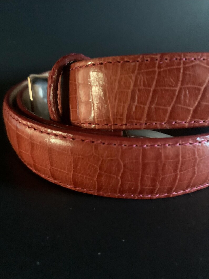 May include: A close-up shot of a brown leather belt with a crocodile skin effect. The belt has a silver buckle and is stitched with a contrasting thread. The leather is a reddish-brown colour.
