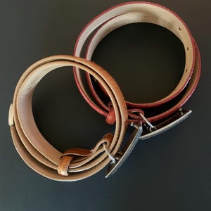 May include: Two leather belts with silver buckles. One is light brown with a textured pattern, the other is red. The belts are coiled on a dark surface.