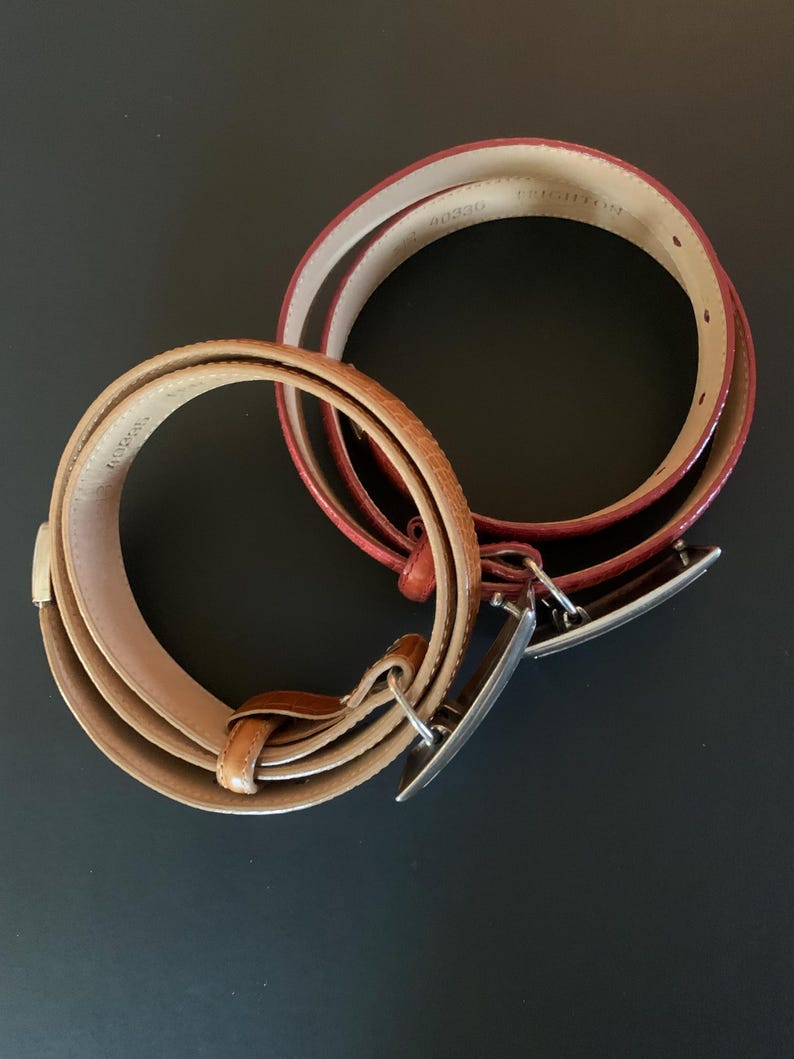 May include: Two leather belts with silver buckles. One is light brown with a textured pattern, the other is red. The belts are coiled on a dark surface.