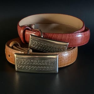 May include: Two leather belts with silver-toned rectangular buckles. One belt is a reddish-brown colour, and the other is tan. Both belts have a textured, embossed pattern. The buckles have a decorative, engraved design.