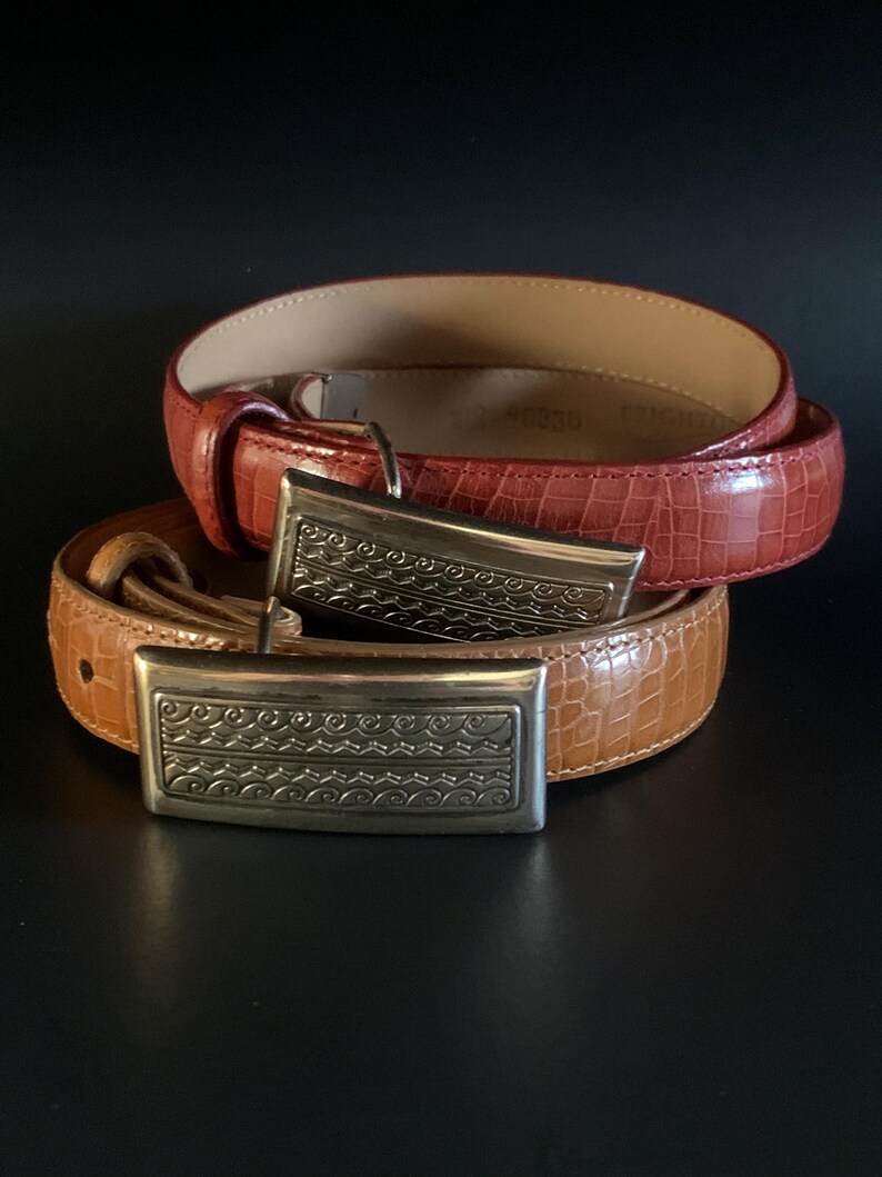 May include: Two leather belts with silver-toned rectangular buckles. One belt is a reddish-brown colour, and the other is tan. Both belts have a textured, embossed pattern. The buckles have a decorative, engraved design.