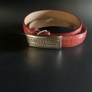 May include: A red leather belt with a silver rectangular buckle. The belt has a textured surface and is coiled on a dark background. The buckle features an ornate design.