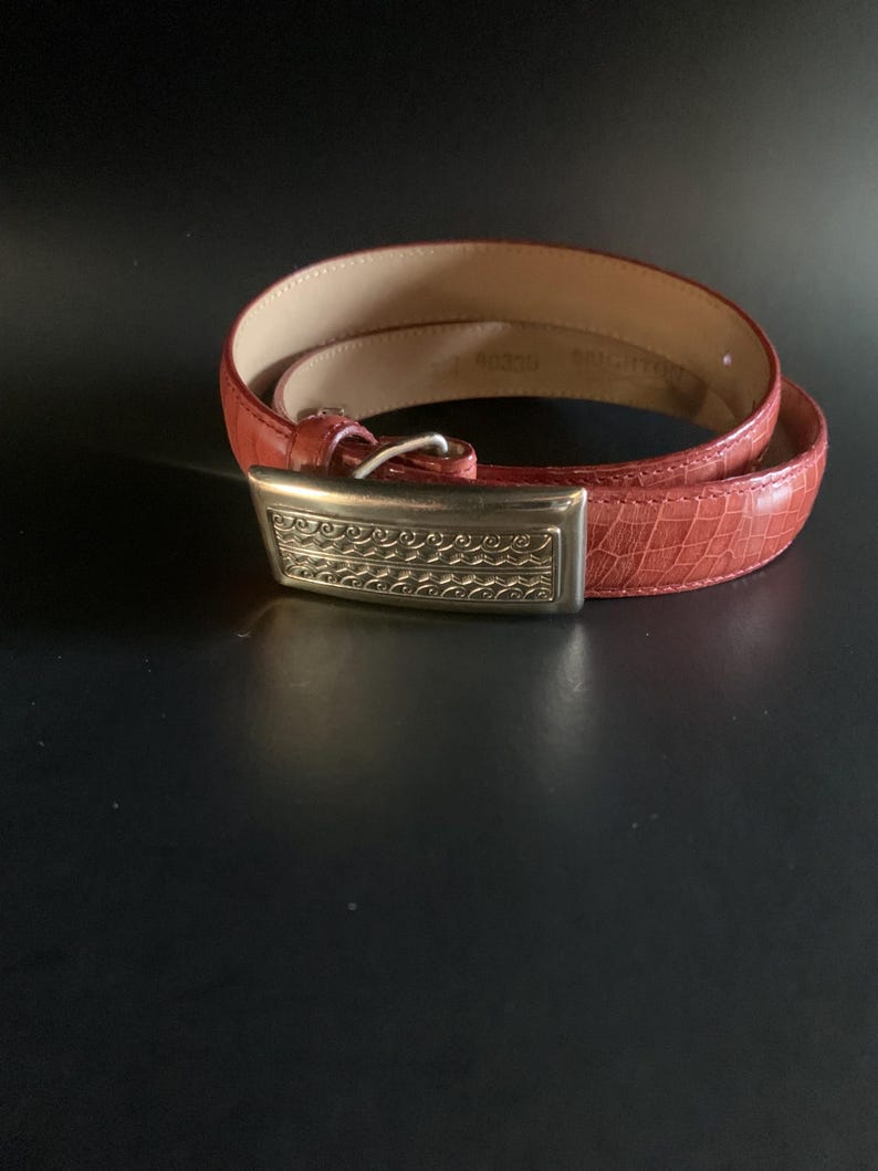 May include: A red leather belt with a silver rectangular buckle. The belt has a textured surface and is coiled on a dark background. The buckle features an ornate design.