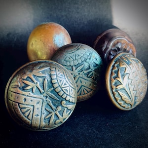 May include: A collection of antique bronze door knobs with intricate embossed designs. The knobs vary in size and shape, showcasing detailed patterns and a weathered patina. The color palette includes shades of bronze, copper, and hints of verdigris.