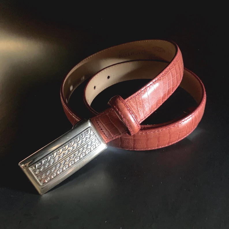 May include: A reddish-brown leather belt with a silver-coloured buckle. The belt has a textured, crocodile-like pattern. The buckle is rectangular with an ornate design. The belt is rolled up on a dark surface.