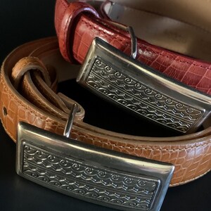 May include: Two leather belts with silver rectangular buckles. One is reddish-brown, the other tan. The buckles feature an engraved design. The belts are arranged in a layered composition. The belts are likely to be measured in centimetres.