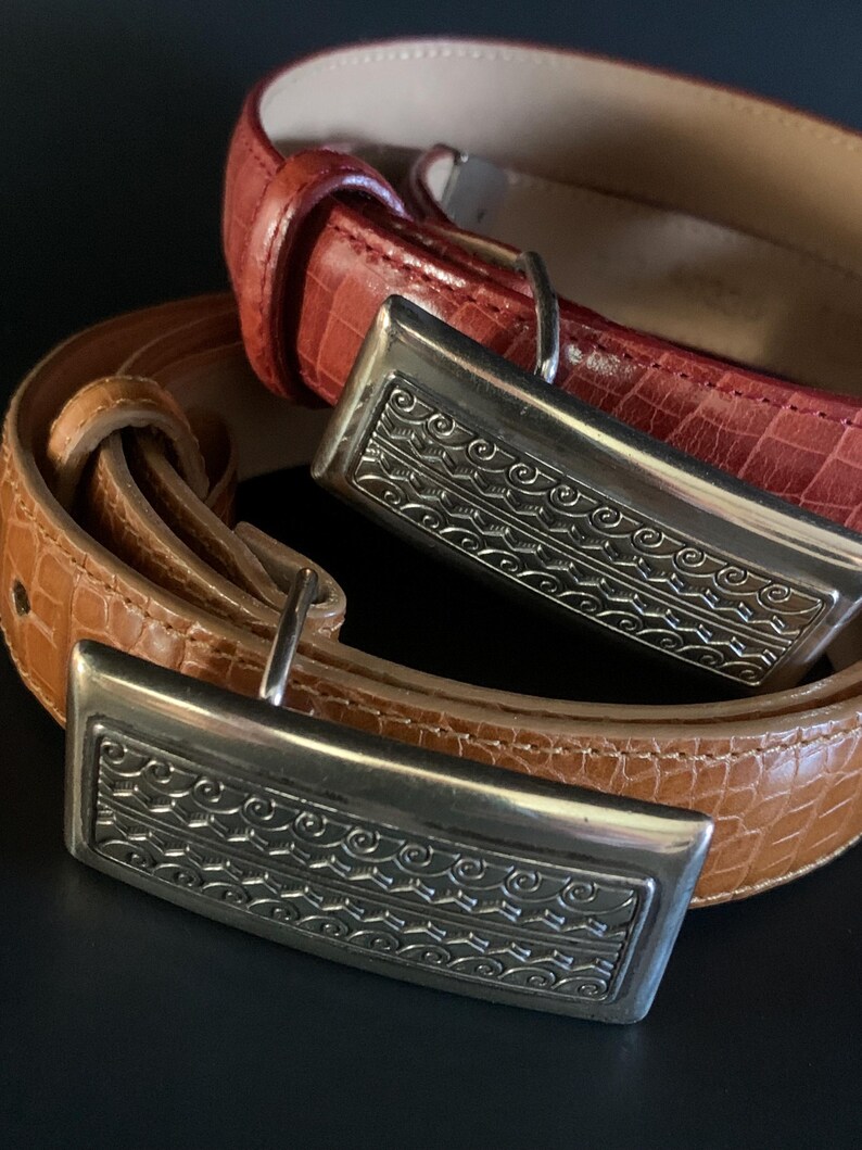 May include: Two leather belts with silver rectangular buckles. One is reddish-brown, the other tan. The buckles feature an engraved design. The belts are arranged in a layered composition. The belts are likely to be measured in centimetres.