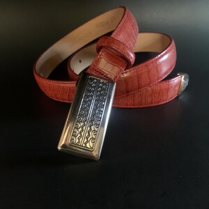 May include: A red belt with a crocodile-effect texture. It has a silver rectangular buckle with an ornate pattern. The belt is coiled on a dark surface, highlighting its colour and texture.