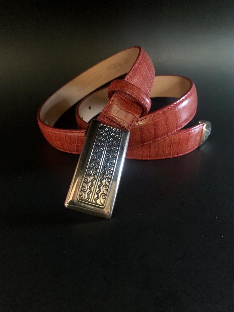 May include: A red belt with a crocodile-effect texture. It has a silver rectangular buckle with an ornate pattern. The belt is coiled on a dark surface, highlighting its colour and texture.
