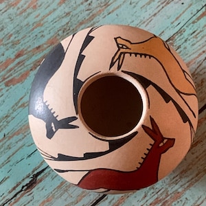May include: A round, beige ceramic pot with a central hole. The pot is decorated with stylized animal figures in black, red, and orange. The design includes a snake, a deer, and abstract shapes. The pot has a rustic, handcrafted appearance.