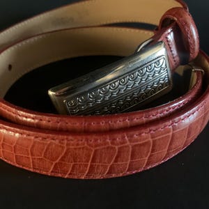 May include: A red belt with a silver buckle. The belt has a textured, crocodile-like pattern. The silver buckle has an ornate, engraved design. The inside of the belt is a light tan colour.