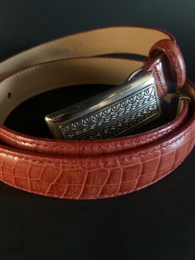 May include: A red belt with a silver buckle. The belt has a textured, crocodile-like pattern. The silver buckle has an ornate, engraved design. The inside of the belt is a light tan colour.